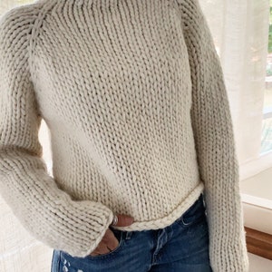 May include: A cream-colored knitted sweater with a mock neck and long sleeves. The sweater has a chunky knit texture and a ribbed hem.