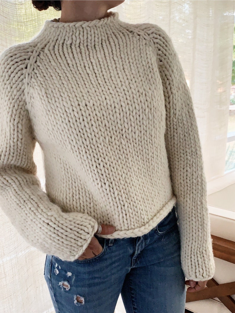 May include: A cream-colored knitted sweater with a mock neck and long sleeves. The sweater has a chunky knit texture and a ribbed hem.