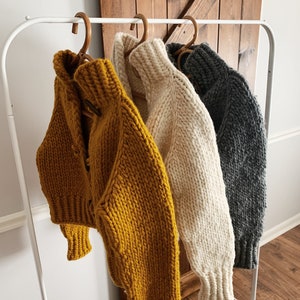 以下が含まれることがあります： 白いラックにかけられた、厚手のニットセーター3枚。セーターはマスタードイエロー、クリーム、グレーです。