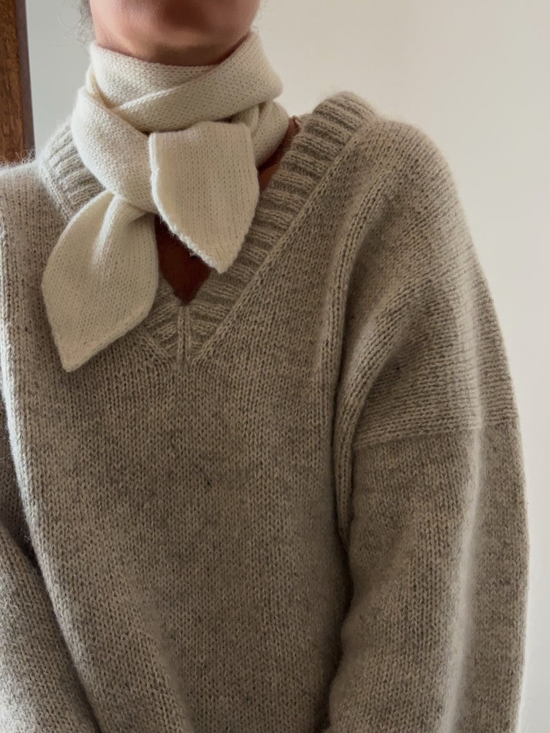 woman standing, wearing a cozy v neck sweater in light gray color and a white thin scarf about 7.5 cm wide, wrapped twice around her neck and tied in one simple knot. Scarf is pointy on both ends.