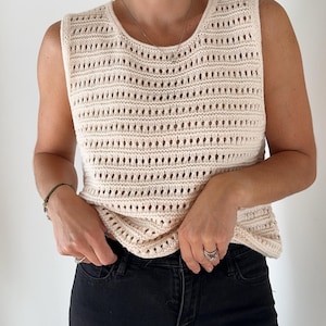 May include: A cream-colored crochet sleeveless top with a delicate open-weave pattern. The top is worn with black jeans.