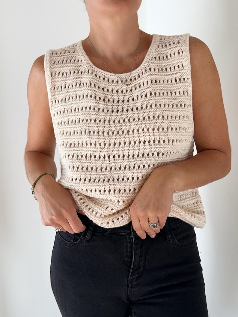 May include: A cream-colored crochet sleeveless top with a delicate open-weave pattern. The top is worn with black jeans.