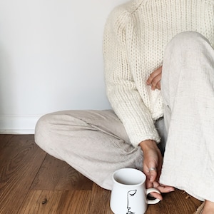 May include: A white ceramic mug with a black line drawing of a face on it. The mug is sitting on a wooden floor. A person is sitting on the floor with their legs crossed, wearing a white sweater and beige pants.