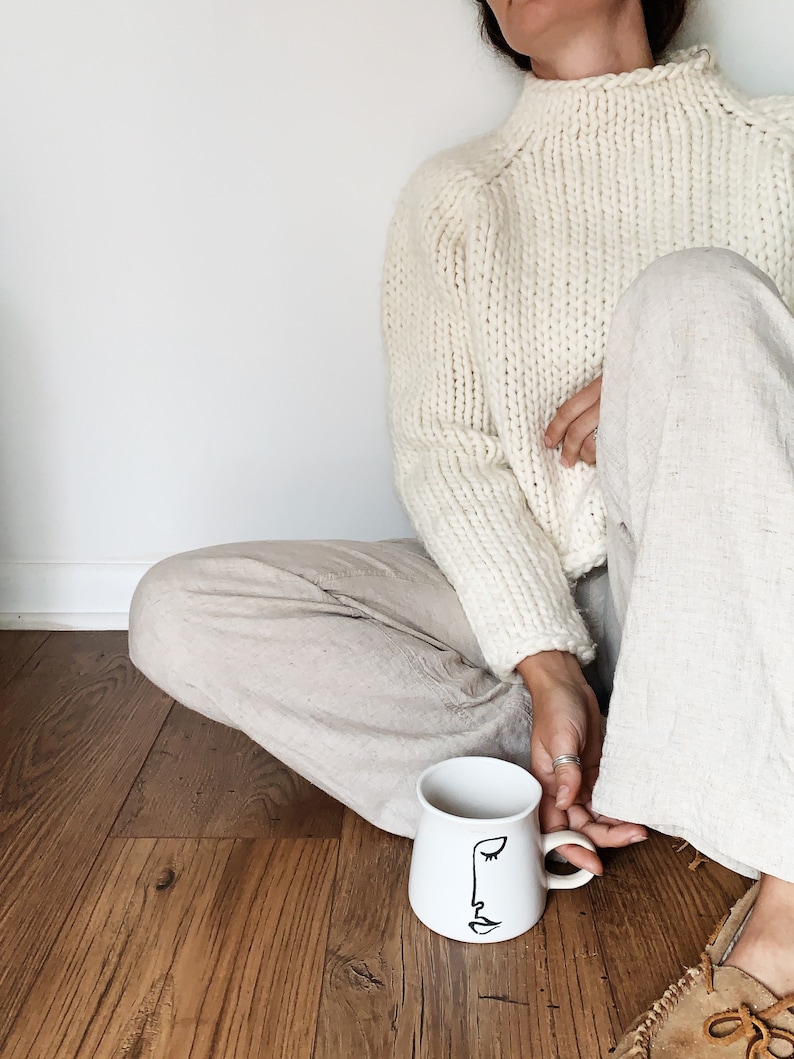 May include: A white ceramic mug with a black line drawing of a face on it. The mug is sitting on a wooden floor. A person is sitting on the floor with their legs crossed, wearing a white sweater and beige pants.