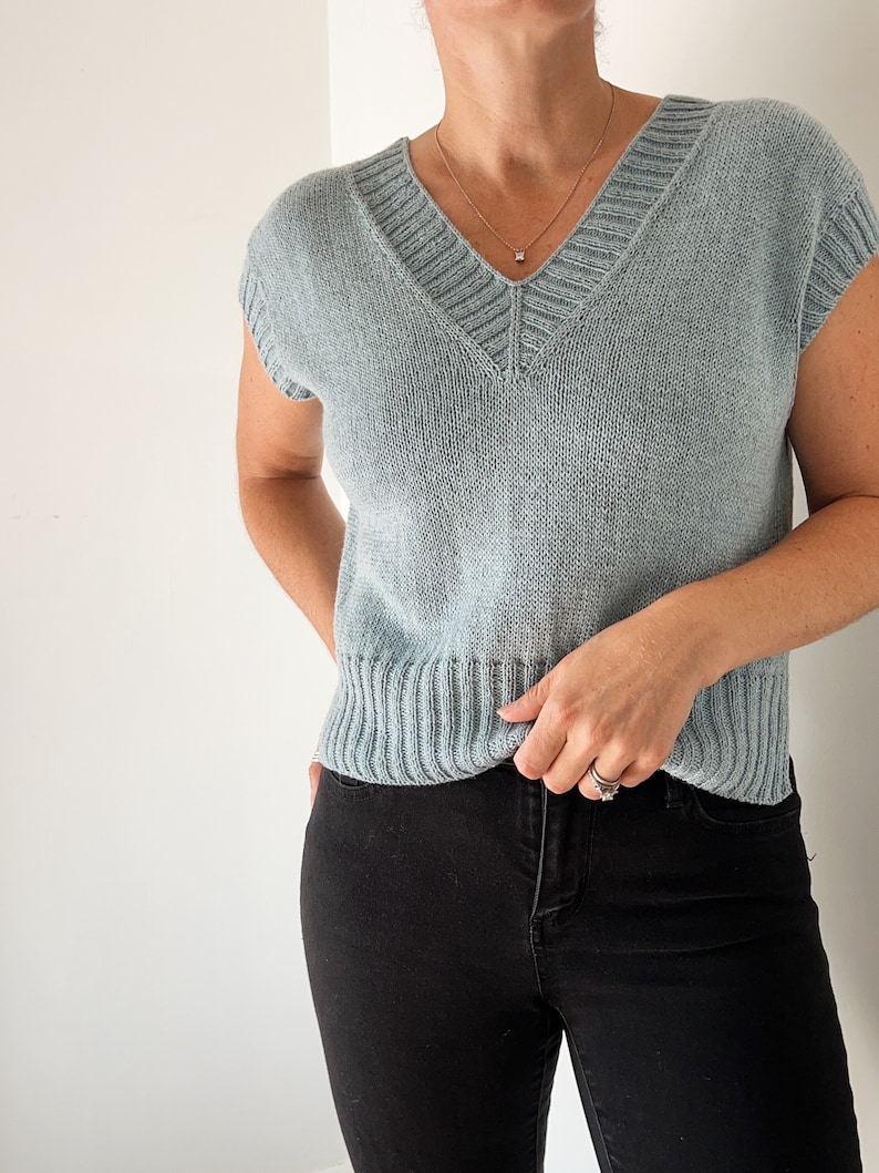 May include: A light blue sleeveless knitted sweater with a v-neck and a ribbed hem. The sweater is worn over black jeans.