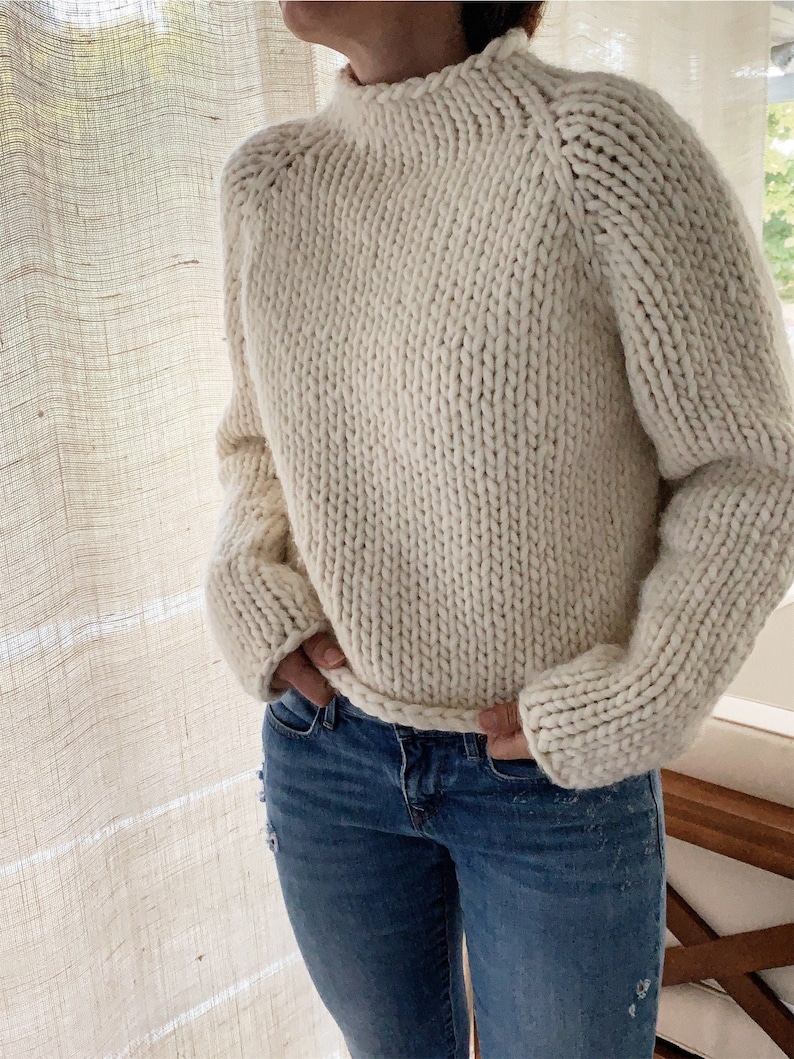 May include: A cream-colored knitted sweater with a high neck and long sleeves. The sweater is made of a thick yarn and has a relaxed fit.