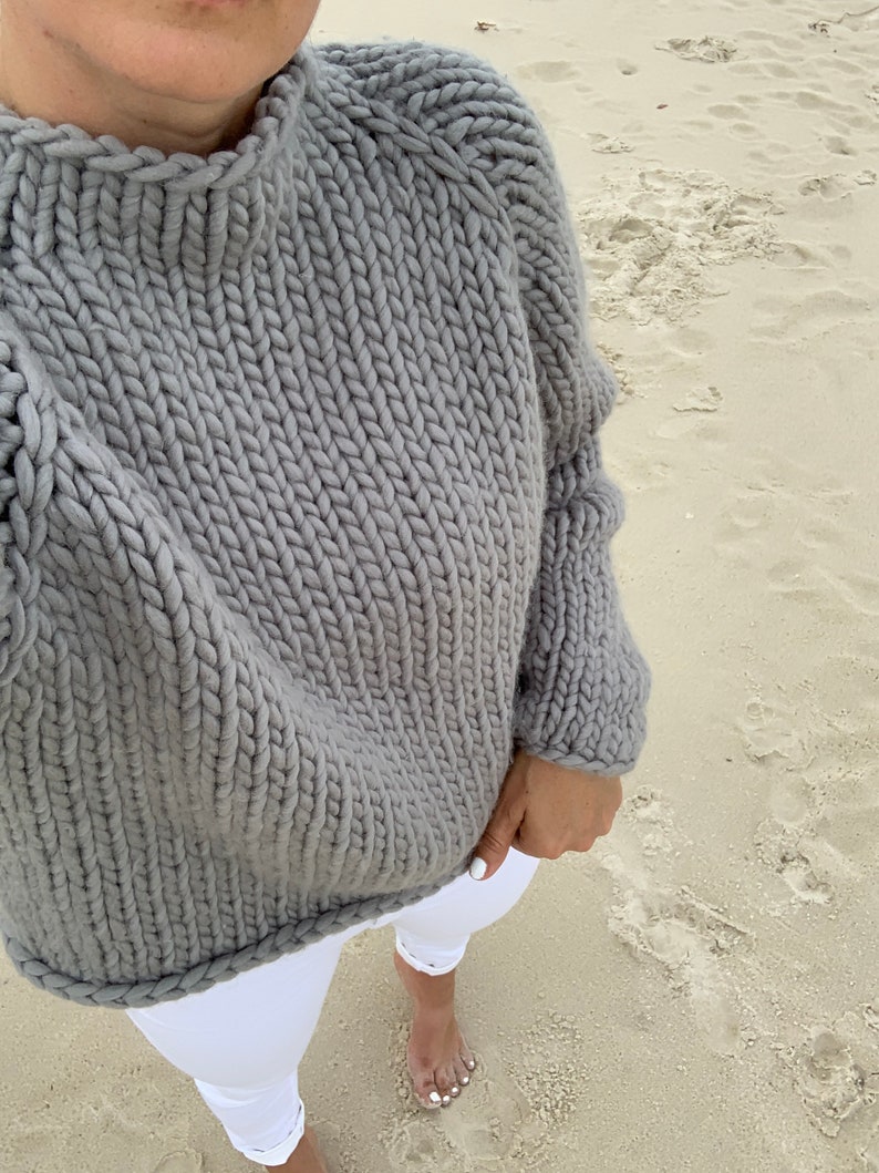 May include: A gray chunky knit sweater with a high neckline and long sleeves. The sweater is made of a thick yarn and has a relaxed fit.