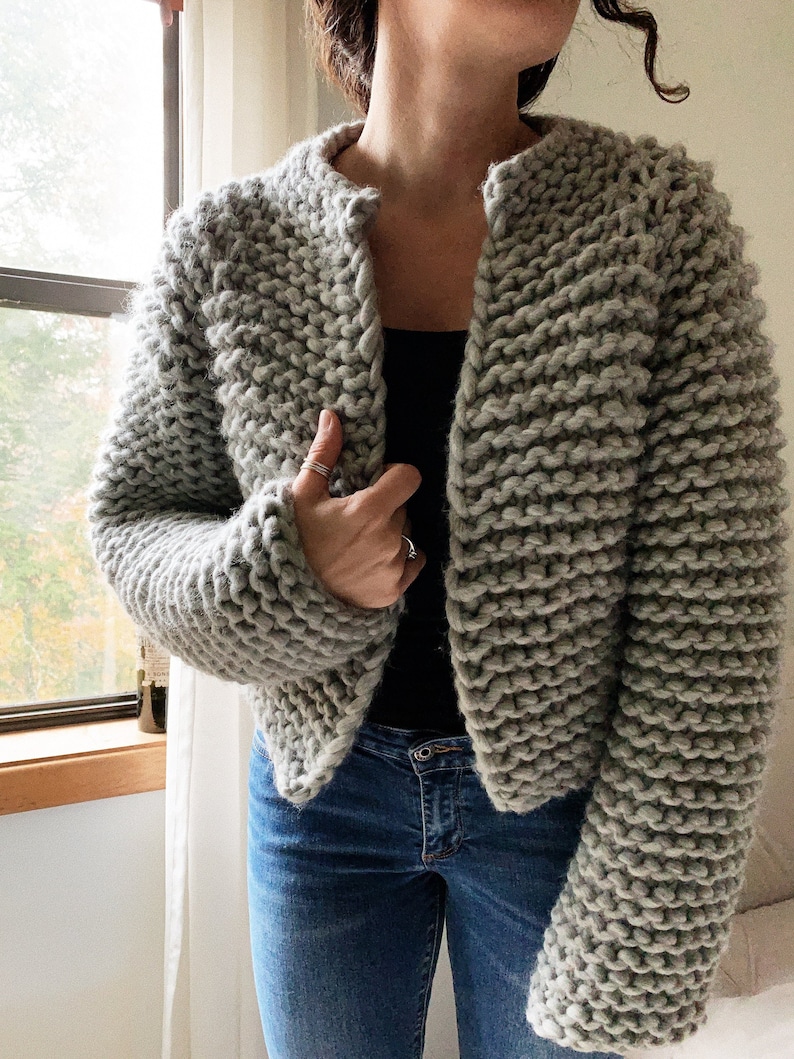 K&ouml;nnte beinhalten: Ein grauer Strickcardigan mit leicht verk&uuml;rzter L&auml;nge und langen &Auml;rmeln. Der Cardigan ist aus einem dicken Garn gefertigt und hat ein schlichtes, klassisches Design.