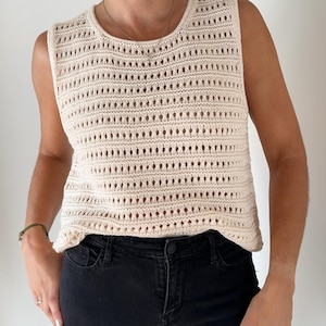 May include: A cream-colored crochet tank top with a delicate open-weave pattern. The top is sleeveless and has a round neckline.