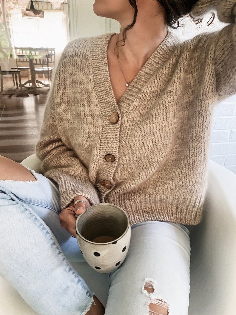 May include: A woman wearing a light brown knit cardigan with wooden buttons and light blue jeans is holding a white ceramic mug with black polka dots.