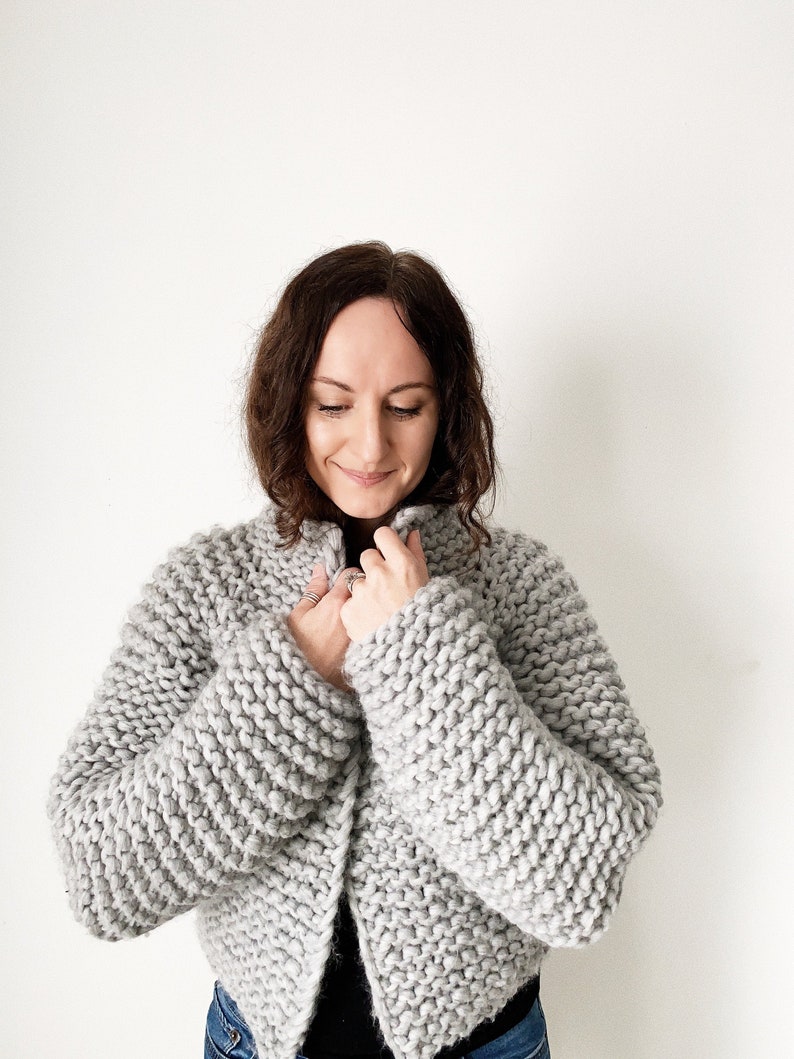 K&ouml;nnte beinhalten: Ein grauer, kurzer Cardigan mit langen &Auml;rmeln und leicht offenem Vorderteil. Der Cardigan ist aus einem dicken Strickgarn gefertigt.