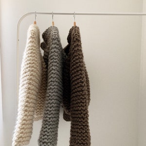 K&ouml;nnte beinhalten: Drei Strickpullover h&auml;ngen an einem wei&szlig;en Gestell. Die Pullover sind in den Farben Beige, Grau und Braun.