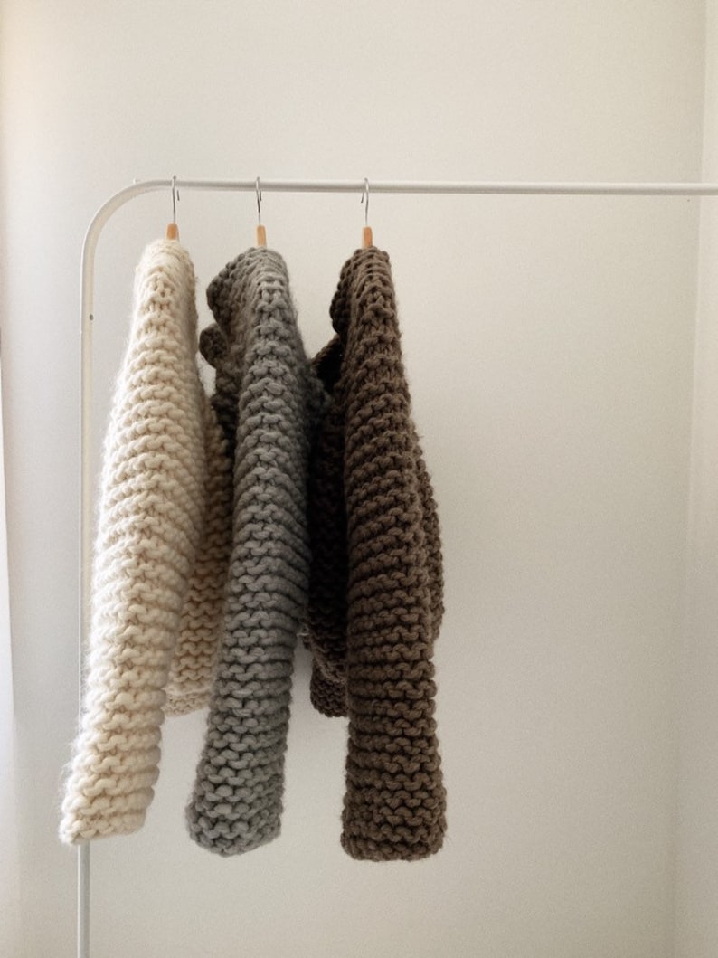 K&ouml;nnte beinhalten: Drei Strickpullover h&auml;ngen an einem wei&szlig;en Gestell. Die Pullover sind in den Farben Beige, Grau und Braun.