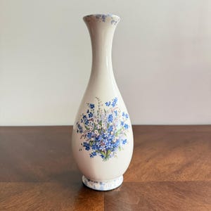 May include: A cream-colored ceramic bud vase with a slender neck and a flared rim. The body features a central floral motif of blue forget-me-nots and small white flowers with green stems. The rim and base have a mottled blue and white pattern.