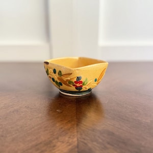 May include: A small, square, yellow ceramic bowl with hand-painted floral and olive branch designs. The bowl features a decorative rim with green, blue, red, and brown accents. The bowl is likely for serving dips or small portions.