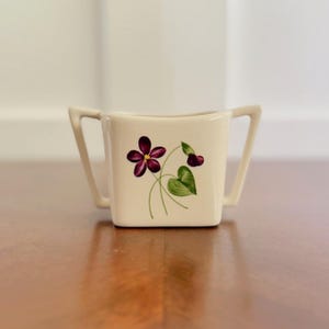 May include: Cream-colored, square sugar bowl with two handles. The front features a floral design with a deep purple flower, buds, and green leaves. The bowl sits on a wooden surface.