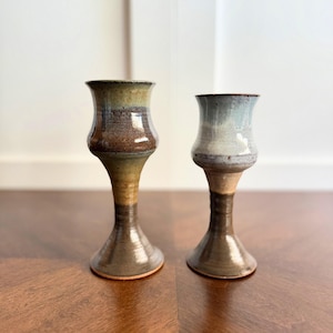 May include: Two handmade ceramic candle holders. The candle holders have a flared top, a bulbous midsection, and a flared base. The candle holders are glazed in shades of brown, tan, and blue. The candle holders are displayed on a wooden surface.