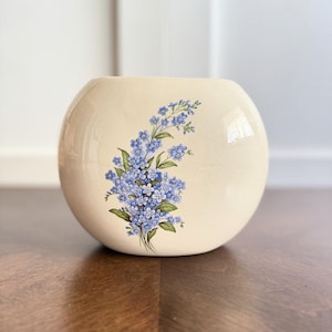 May include: Cream-colored, round ceramic vase with a floral design. The vase features a cluster of blue forget-me-nots with green leaves. The vase is sitting on a wooden surface.