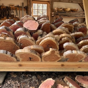 May include: A large pile of freshly cut, irregular wood slices with reddish-brown centers and light-colored edges. The wood pieces are stacked on a wooden shelf in a workshop setting, suggesting a craft or woodworking project.