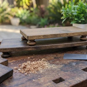 May include: Two rectangular wooden risers with a dark brown stained finish, each featuring four rounded, turned feet. The risers are positioned on a workbench surface alongside wood shavings and a metal hand saw blade, showcasing the wood grain.