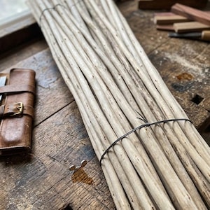 May include: A bundle of light-colored, slender wooden sticks tied together with black wire. The sticks have a natural, weathered appearance. The bundle rests on a rustic wooden surface, with a leather case and tools in the background.