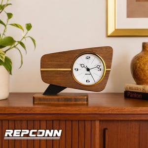 Puede incluir: Un reloj de escritorio de madera con marco trapezoidal, esfera blanca circular con borde dorado y manecillas negras. El reloj presenta el texto REPCONN en la esfera, una línea dorada horizontal y una pieza de soporte negra sobre una base de madera.