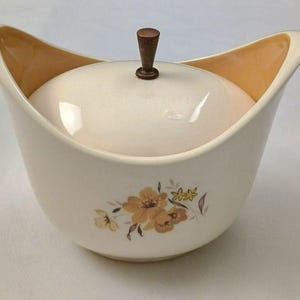 May include: A cream-colored ceramic sugar bowl with a matching lid and a dark brown knob handle. The bowl features high, curved sides and a floral decal in shades of yellow and brown on the front. The interior of the bowl is a soft tan color.