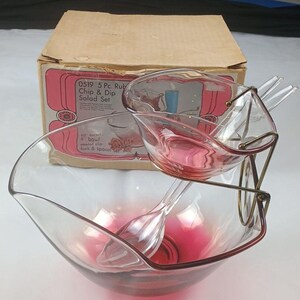 May include: A 5-piece chip and dip salad set featuring a large clear glass bowl with a pink-tinted base, a smaller matching bowl, a clear plastic fork and spoon, and a gold-tone metal stand. The original cardboard box is labeled 0519 5 Pc. Rub.