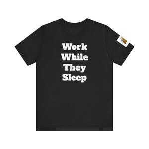 May include: Black t-shirt with the white text "Work While They Sleep" in a bold, sans-serif font. The shirt has a short sleeve design and a small graphic on the right sleeve.