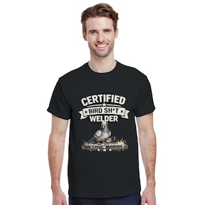 May include: Black t-shirt featuring the text "CERTIFIED BIRD SH*T WELDER" above an illustration of a pigeon on a metal beam with welding sparks. The shirt is made of a soft material.
