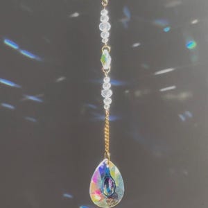 Puede incluir: Un atrapasoles de cristal con una cadena dorada y cuentas transparentes. El cristal en forma de lágrima refracta la luz, mostrando un arcoíris de colores. El atrapasoles cuelga sobre un fondo oscuro.