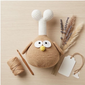May include: A handcrafted crochet owl toy in tan, white, and yellow. The owl features large white eyes, a yellow beak, and a white head. A ball of twine, a crochet hook, and a blank tag are also present.