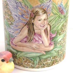 May include: A ceramic mug with a pink rim and a colourful illustration of a fairy with wings sitting on a leafy plant. The fairy has brown hair, a pink dress, and a flower in her hair.