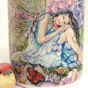 May include: A ceramic mug with a turquoise rim features a watercolour illustration of a fairy with blue wings and a blue dress sitting amongst purple flowers and a butterfly.