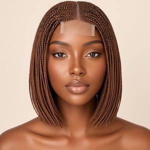 May include: A shoulder-length bob wig featuring fine, uniform micro-braids in a warm chestnut brown colour. The wig includes a sheer lace front panel at the centre part, designed to mimic a natural hairline with individual braided strands.