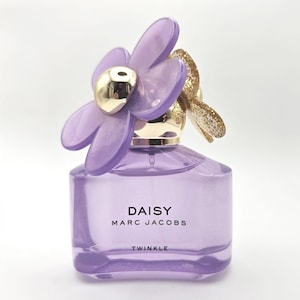 May include: A perfume bottle of Daisy by Marc Jacobs. The bottle is a light purple color with a flower-shaped cap. The flower petals are a translucent purple, and the center is gold. The text "DAISY MARC JACOBS TWINKLE" is printed on the front.