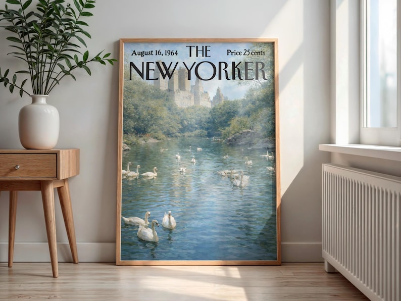 May include: A framed vintage New Yorker magazine cover from August 16, 1964, featuring a serene lake scene with swans and a castle in the background. The artwork is in a wooden frame, displayed indoors near a window and a side table.