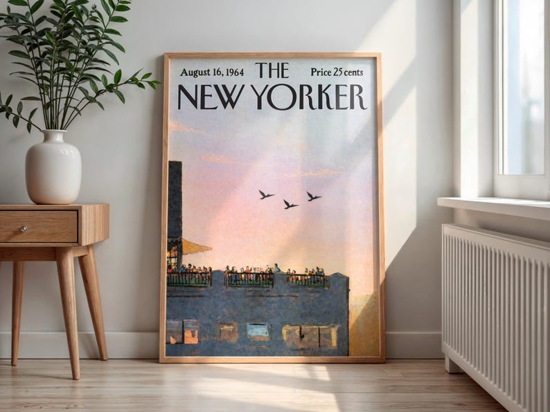 May include: A framed vintage New Yorker magazine cover from August 16, 1964, featuring a sunset scene with birds in flight. The artwork is in a wooden frame, displayed in a well-lit room with a side table and a vase.