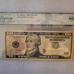 May include: A ten-dollar Federal Reserve Note from 2009, featuring a portrait of Alexander Hamilton. The note is graded and encased in a protective sleeve. The text "We the People" is visible on the note.