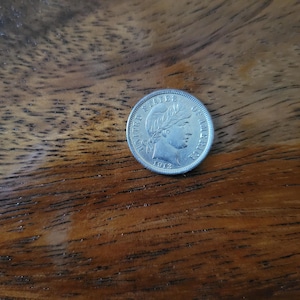 May include: A silver coin from 1912, featuring a profile of a woman and the words "UNITED STATES OF AMERICA." The coin is set against a dark wooden surface, showcasing its intricate details and historical significance.