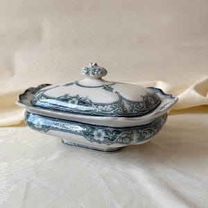 May include: A rectangular ceramic tureen with a lid, featuring a cream-colored base and a dark green floral and scrollwork pattern. The lid has a decorative knob handle, and the edges of both the lid and the base have a scalloped design.