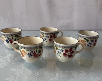 May include: Five ceramic coffee cups with handles. The cups are off-white with a floral design in red, blue, and orange. The rims are decorated with a blue and gold pattern. The cups are arranged on a reflective surface.