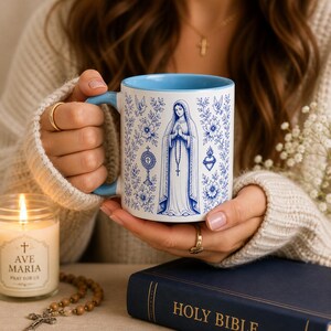 May include: White ceramic mug with a blue interior and handle, decorated with blue floral and religious illustrations. The mug is held in hands, with a lit candle and a book with the words "Holy Bible" visible.