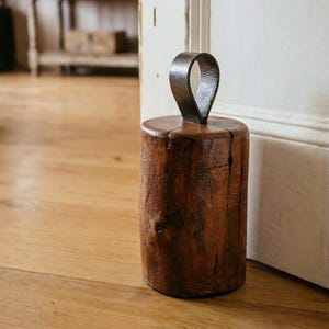 May include: A cylindrical doorstop crafted from a solid piece of dark brown wood with a visible natural grain and texture, featuring a sturdy, arched metal handle attached to the top for easy movement and placement against a door.