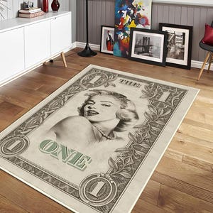 Può includere: Tappeto rettangolare con un design a banconota da un dollaro raffigurante Marilyn Monroe. Il tappeto ha uno sfondo beige con accenti verdi e grigi. La parola "ONE" è visualizzata in verde, ideale per un soggiorno.