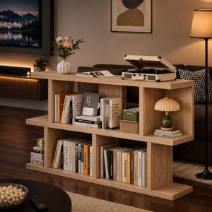 May include: A light oak wood bookshelf with an S-shaped design featuring two horizontal tiers and multiple vertical dividers. The shelves are filled with various books, a cream-colored portable record player, a small green lamp, and a white vase.