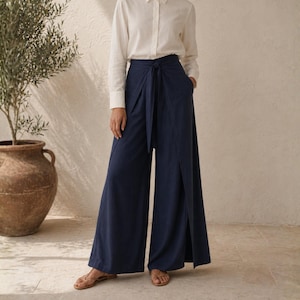 Op de afbeelding: Een witte blouse gecombineerd met een marineblauwe broek met wijde pijpen. De broek heeft een wikkelontwerp met een strik in de taille en zakken. De outfit wordt gecompleteerd met sandalen. Op de achtergrond staat een grote potplant. De broek is ongeveer 100 cm lang.