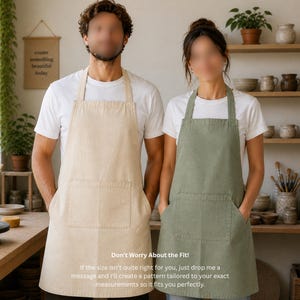 May include: Two people wearing aprons in a pottery studio. One apron is beige, the other is olive green. Both aprons have a large front pocket. The text on the wall says "create something beautiful today". The text at the bottom says "Don't Worry About the Fit!"