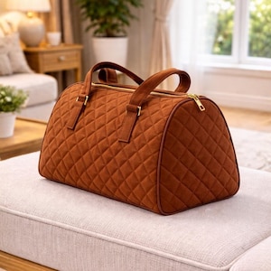 Puede incluir: Una bolsa de lona acolchada marrón con forma redondeada, con cremallera dorada y asas a juego. La bolsa está hecha de un material suave similar al ante y tiene un patrón de diamantes. La bolsa está sobre un banco de color claro.