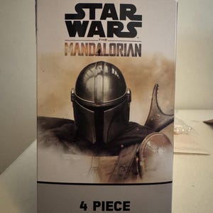 May include: A box for a Star Wars: The Mandalorian full sheet set. The box features the Star Wars logo and the Mandalorian's helmet. The text "4 Piece Full Sheet Set" is printed on the bottom of the box.